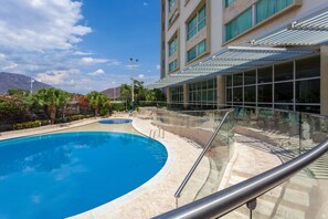 Una piscina con barandilla de vidrio, un edificio con grandes ventanales y palmeras al fondo.