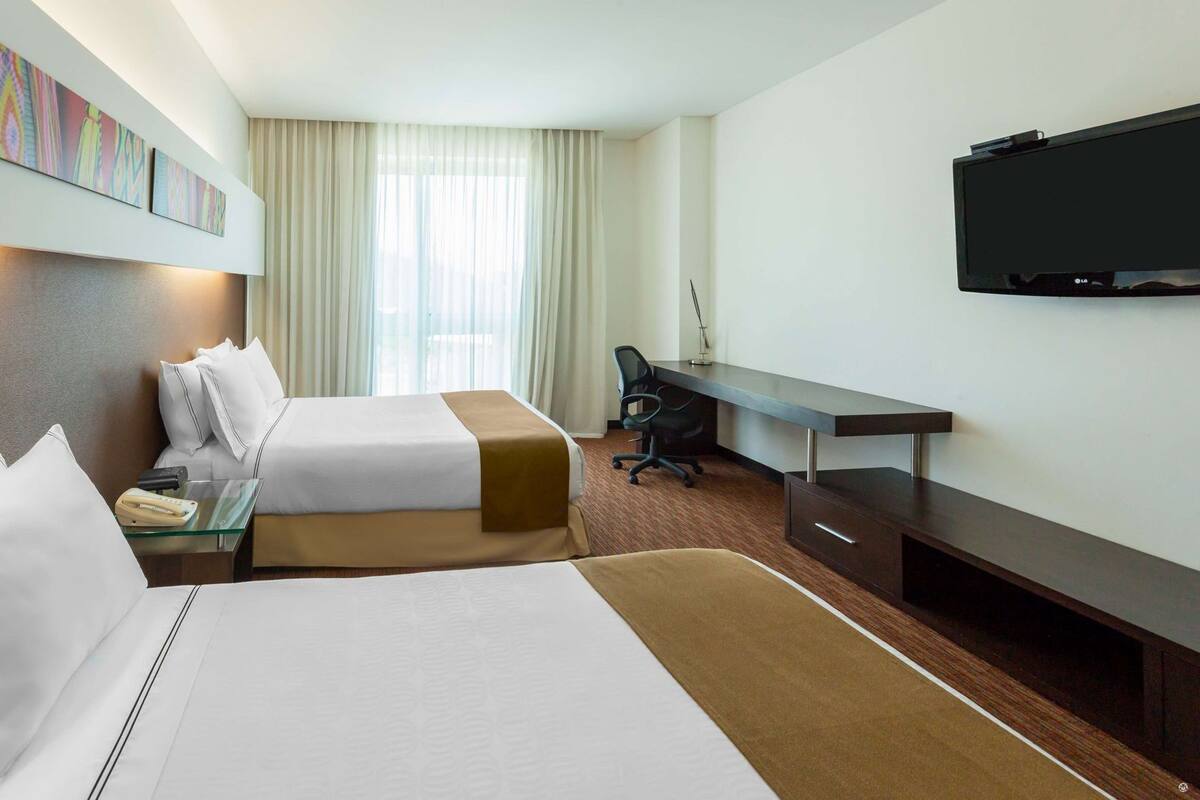 A hotel room with two beds, a desk, a chair, a TV, and a window with curtains.