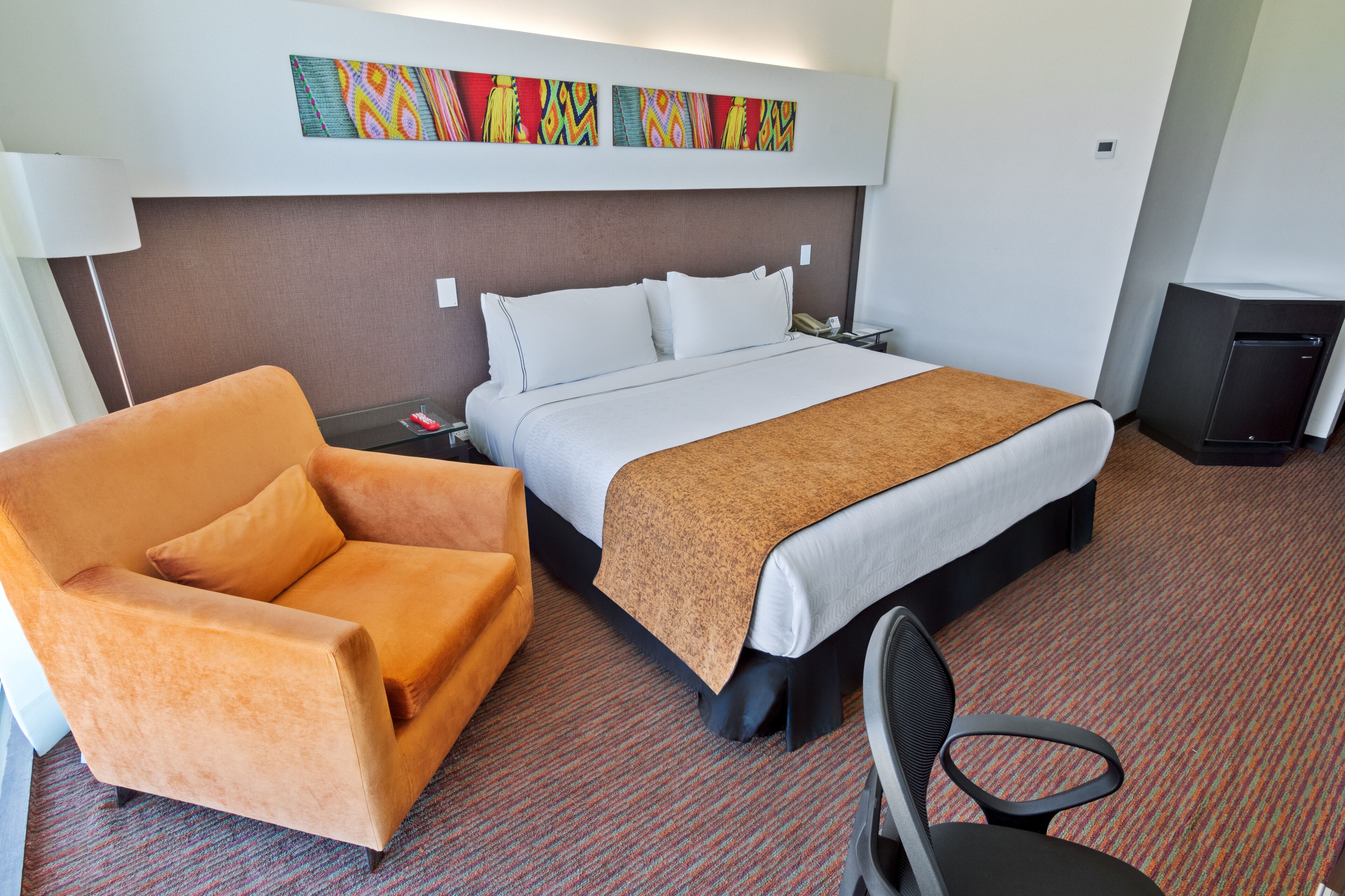 Una habitación de hotel con una cama, un sillón naranja, un escritorio con una lámpara y un televisor pequeño.