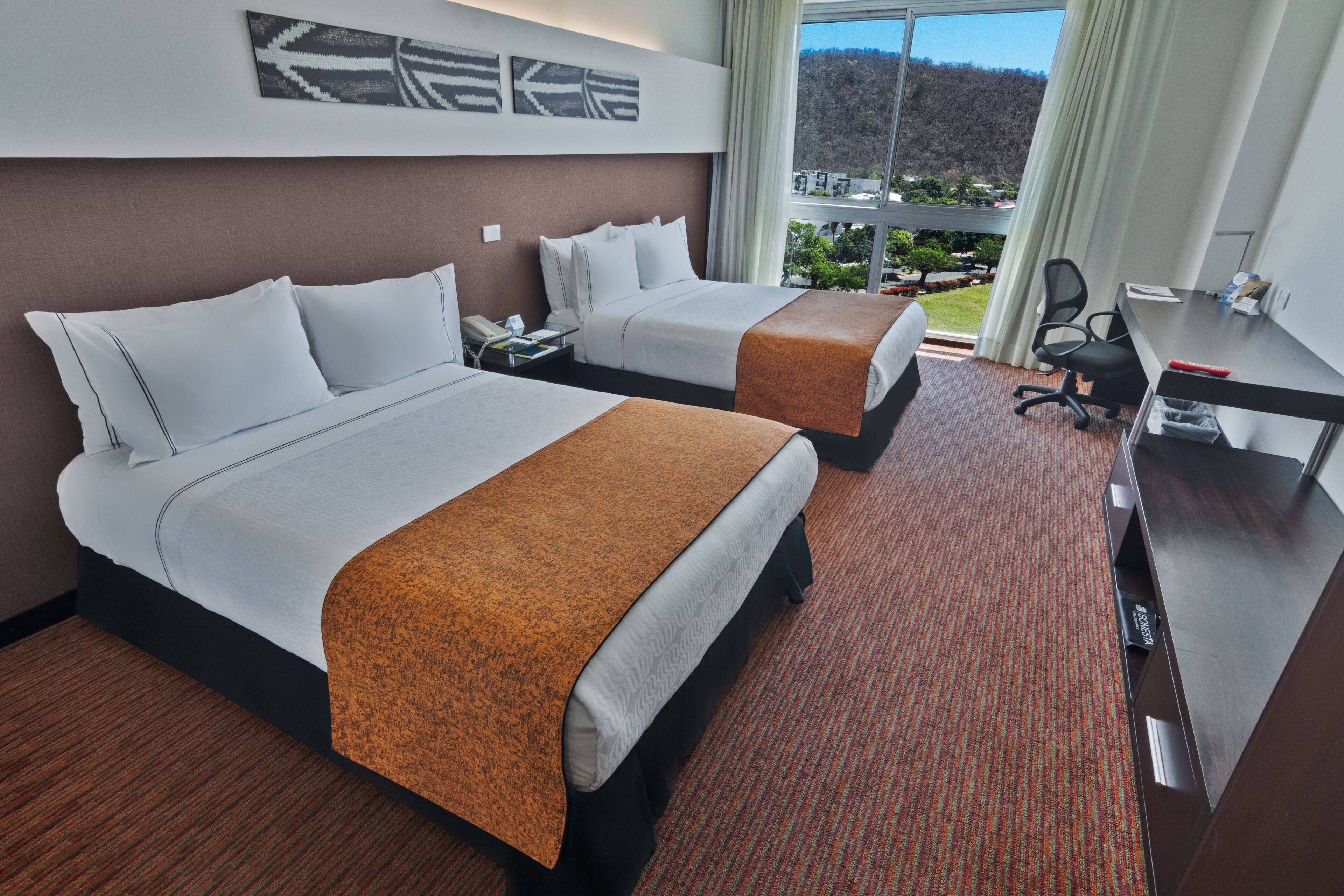 Ein Hotelzimmer mit zwei Betten, einem Schreibtisch und Blick auf eine Berglandschaft.
