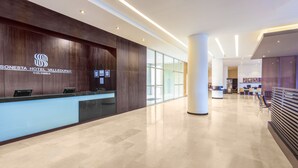 Una moderna zona de recepción de hotel con un mostrador de madera, un muro de cristal y un vestíbulo amplio con techo alto y suelo de color claro.