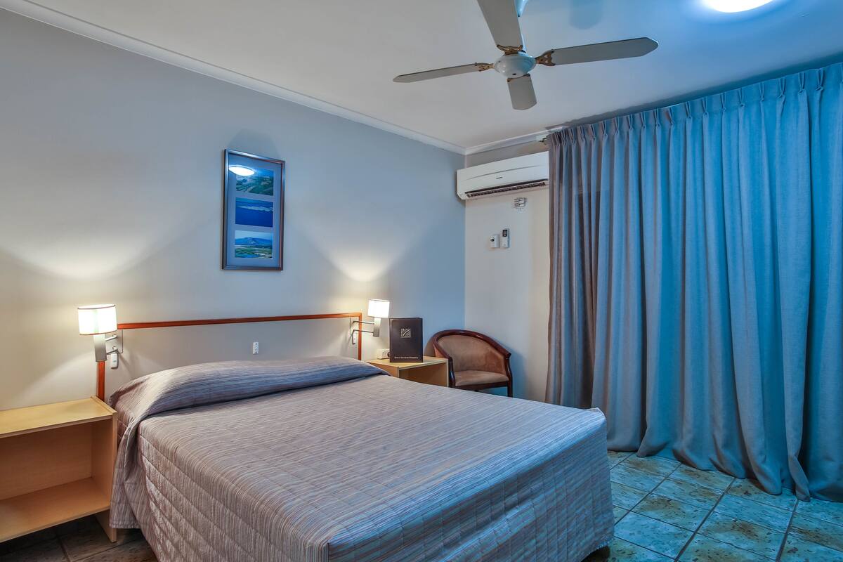 Une chambre d’hôtel comprenant un lit, des tables de chevet, un bureau, une chaise, un ventilateur de plafond et une fenêtre avec des rideaux.