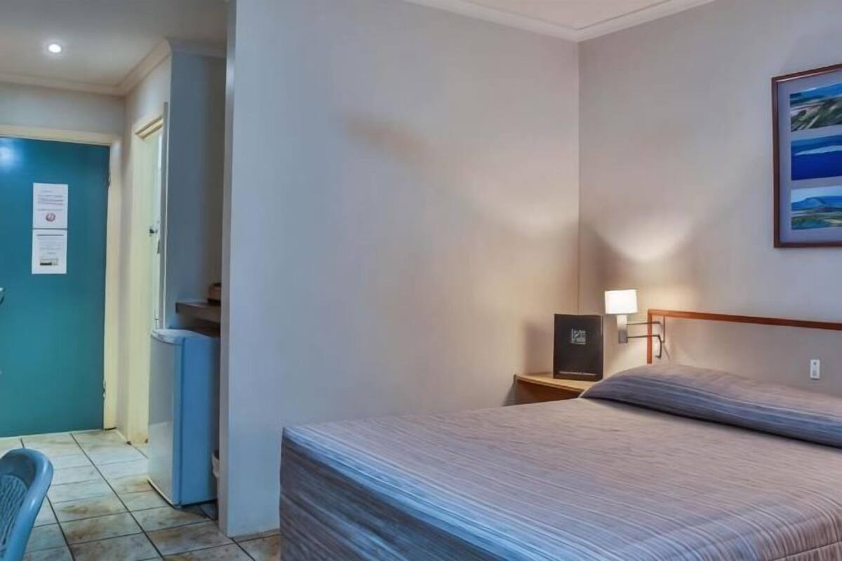Une chambre d’hôtel équipée d’un lit, d’un bureau, d’une chaise, d’une table de chevet avec une lampe et d’un téléviseur fixé au mur.
