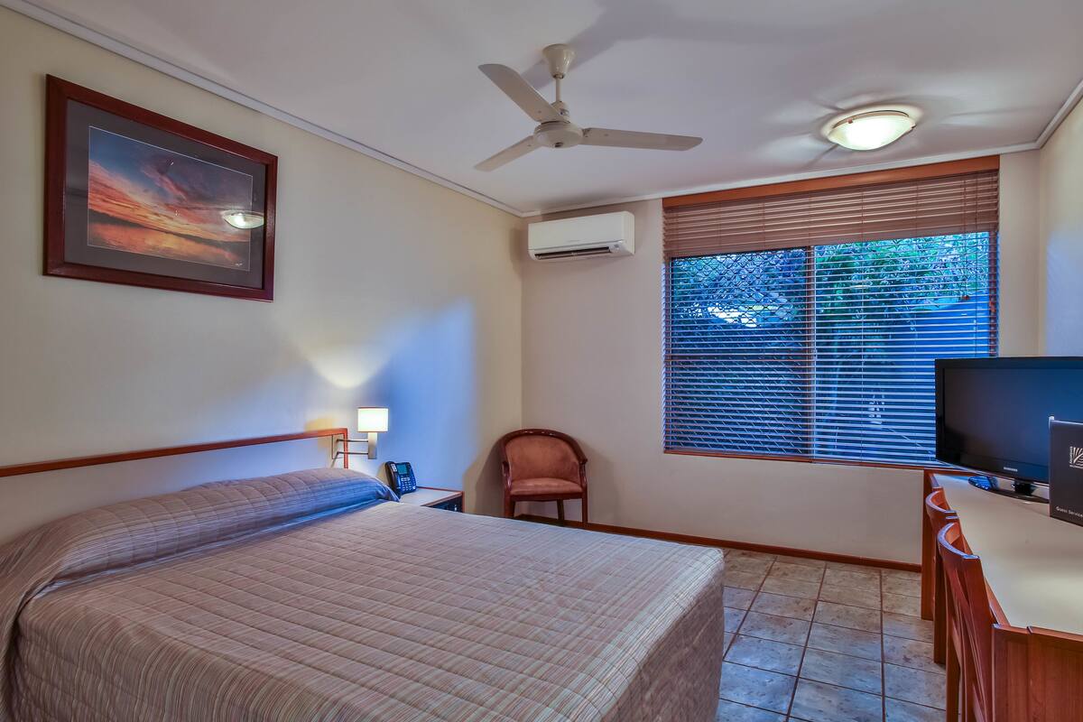 Une chambre d’hôtel comprenant un lit, un bureau avec un ordinateur, une chaise, une fenêtre avec des stores, un climatiseur, un ventilateur de plafond et un tableau encadré au mur.