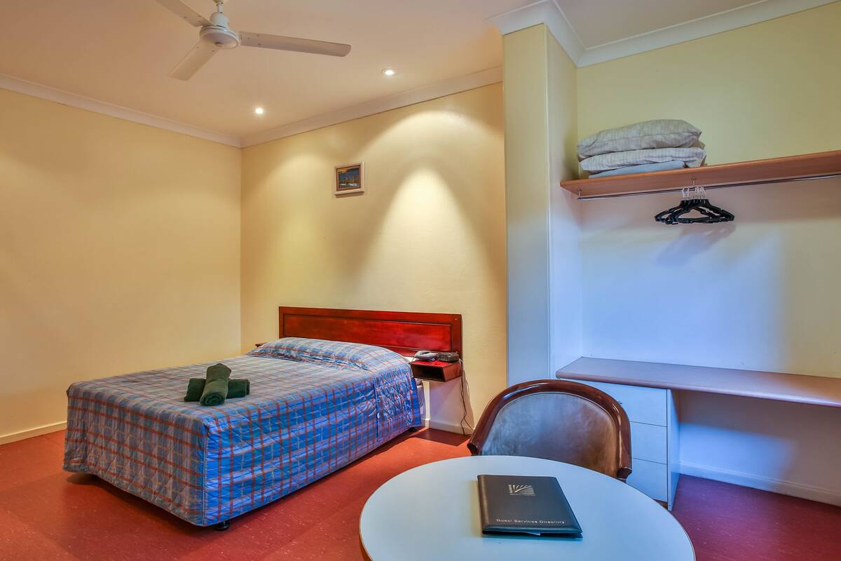 Une chambre d’hôtel comprenant un lit, un bureau, une chaise, une table, un ventilateur de plafond et une étagère avec des serviettes pliées.