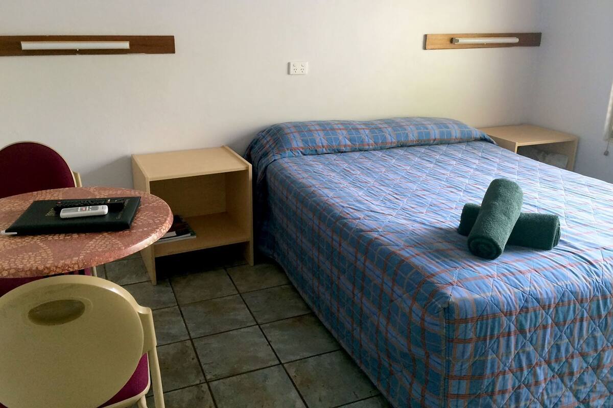 Une petite chambre d’hôtel avec un lit simple, un bureau avec une chaise, une petite table et une étagère.