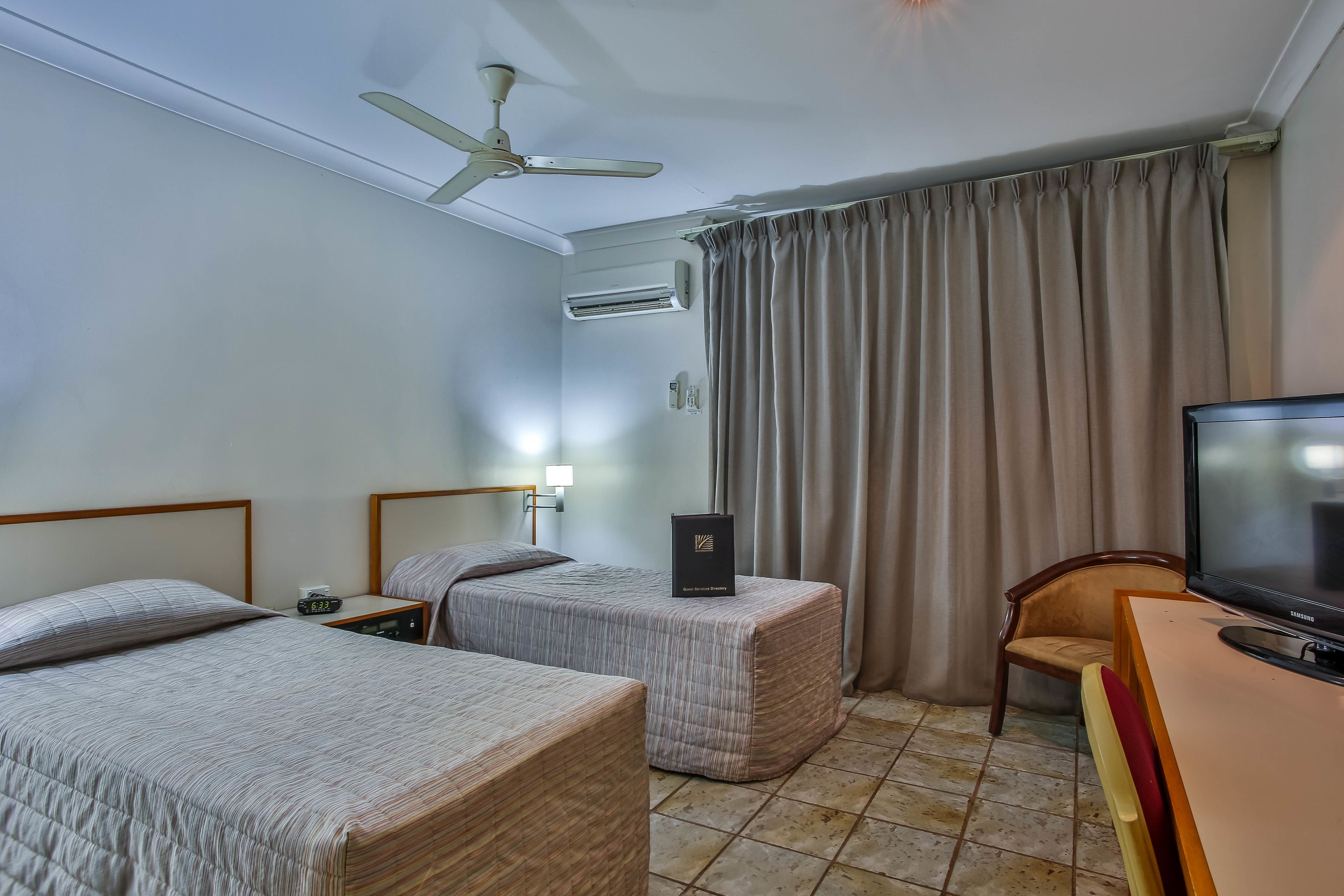A hotel room with two beds, a ceiling fan, an air conditioning unit, a television, and a desk with a chair.