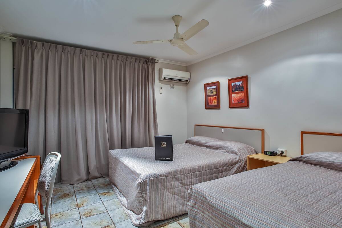 Une chambre d’hôtel avec deux lits, un bureau avec une chaise, un ventilateur de plafond, un appareil de climatisation et deux tableaux encadrés au mur.