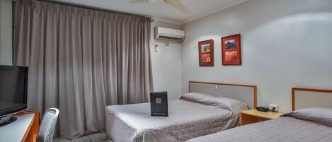 A hotel room with two beds, a desk with a chair, a ceiling fan, an air conditioning unit, and two framed pictures on the wall.