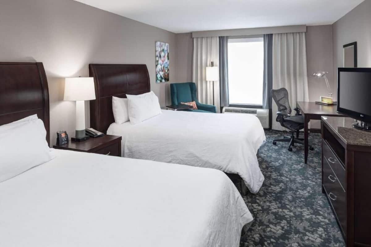 A hotel room with two beds, a desk with a TV, a chair, a lamp, and a window with curtains.