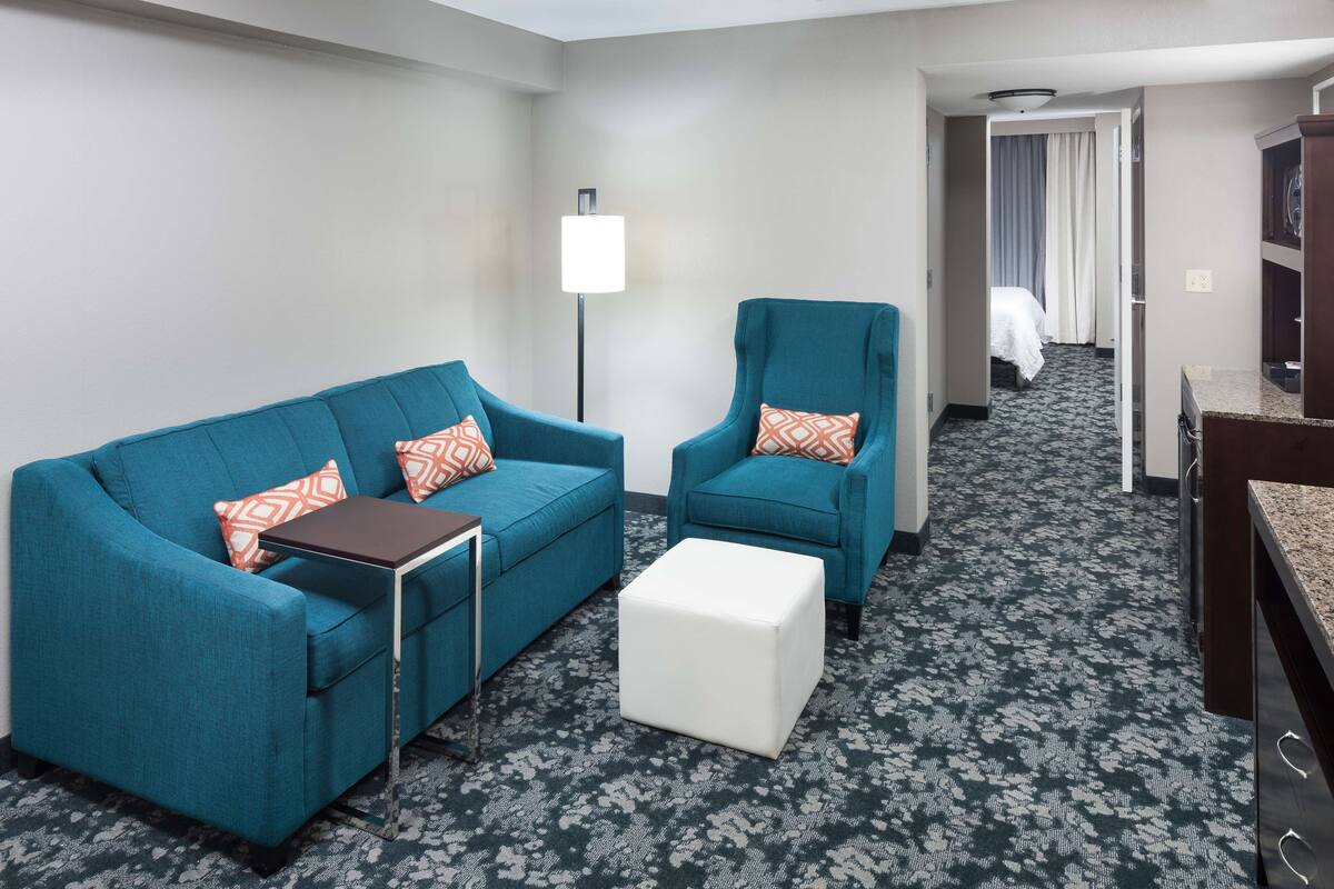 A hotel room with a teal sofa, a matching armchair, a small white ottoman, a side table, and a floor lamp.