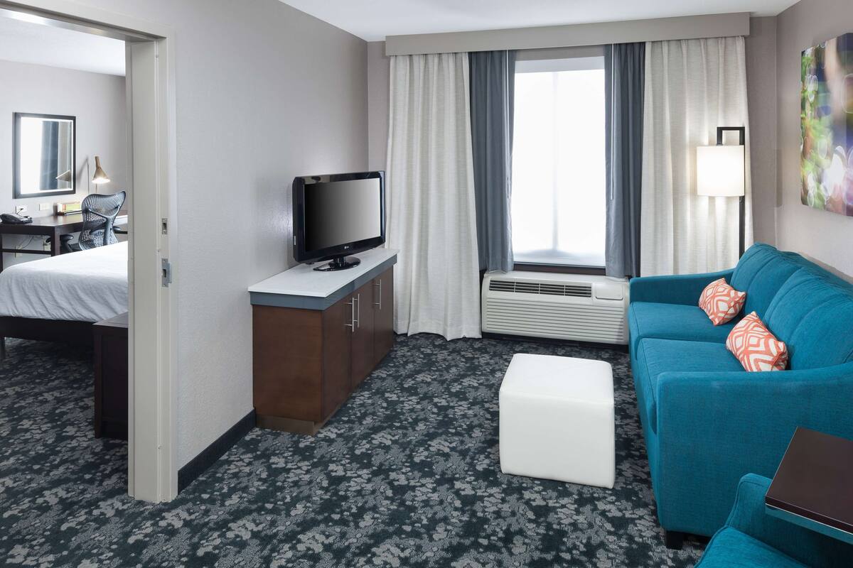 A hotel room with a bed, a desk with a computer, a TV, a sofa, and a window with curtains.
