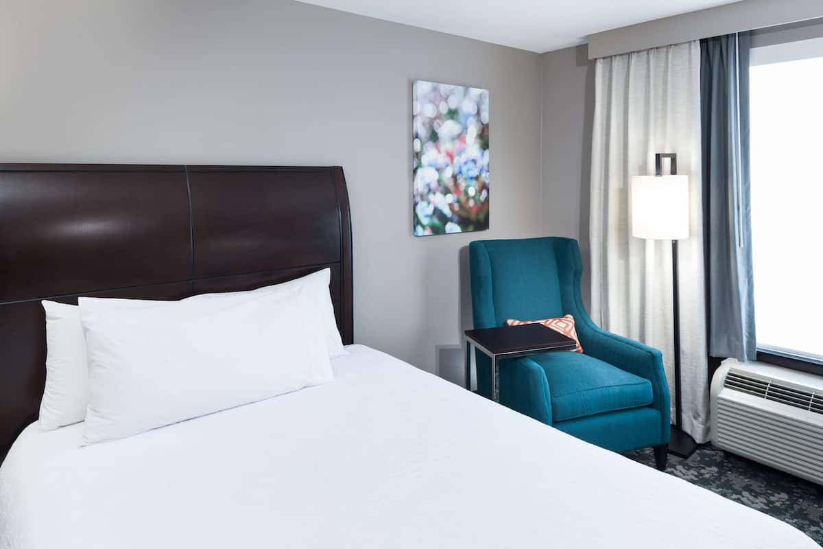A hotel room with a large bed, a teal armchair, a small table, a lamp, and a window with curtains.