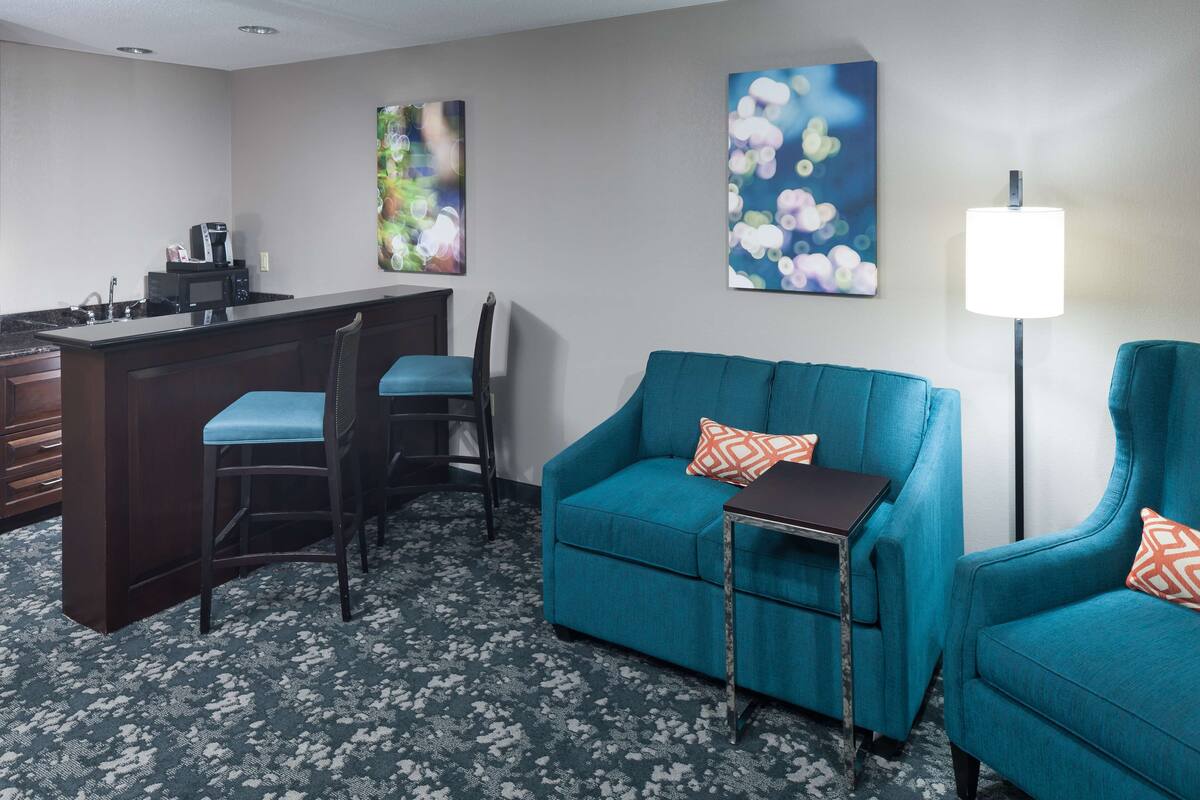 A hotel room with a kitchenette, a teal sofa, two matching armchairs, a small table, and a floor lamp.