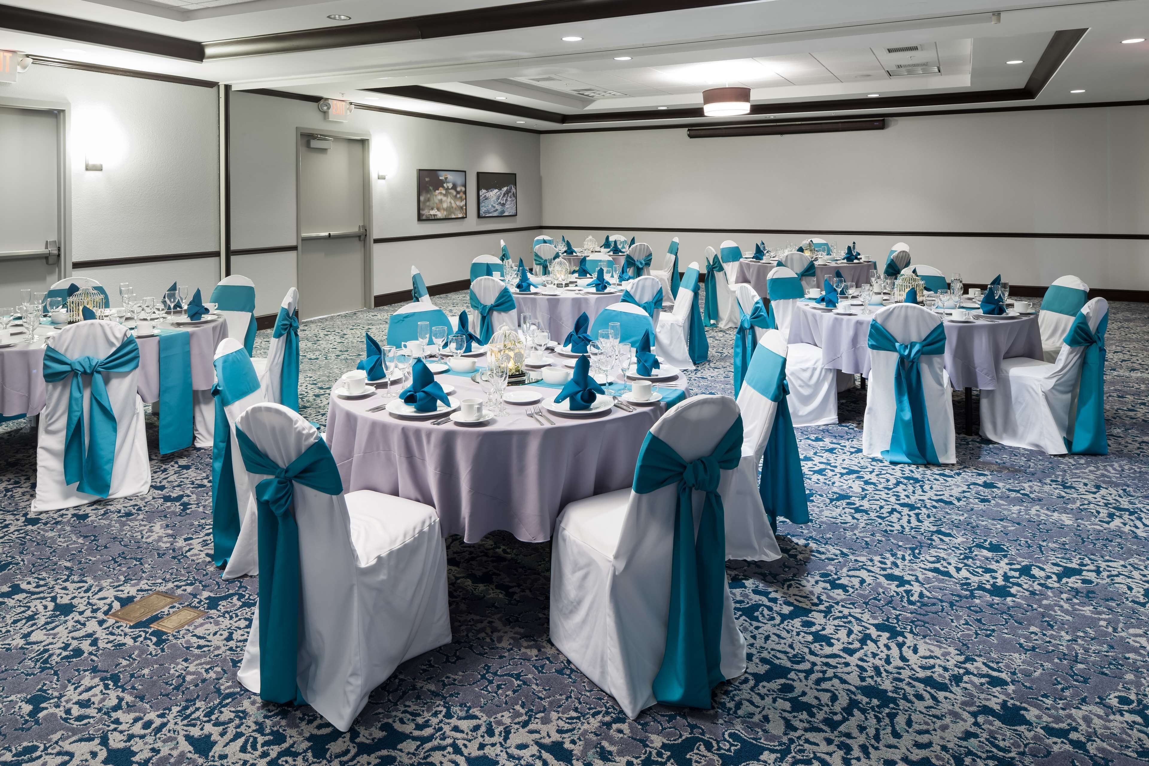 Un salón de conferencias con mesas redondas, sillas blancas con cintas turquesa y alfombra azul.