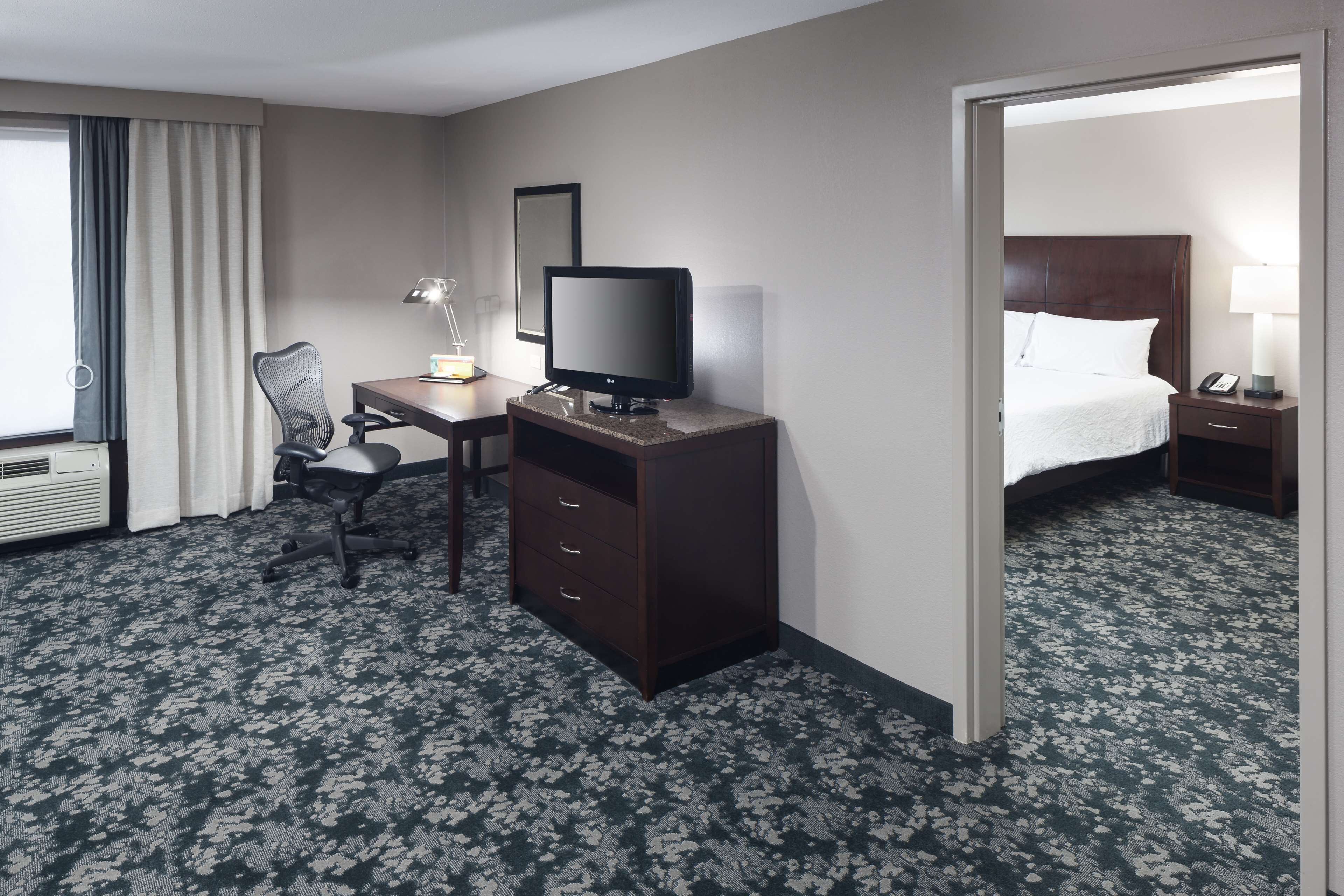 A hotel room with a bed, a desk with a computer, a chair, a TV, and a lamp.