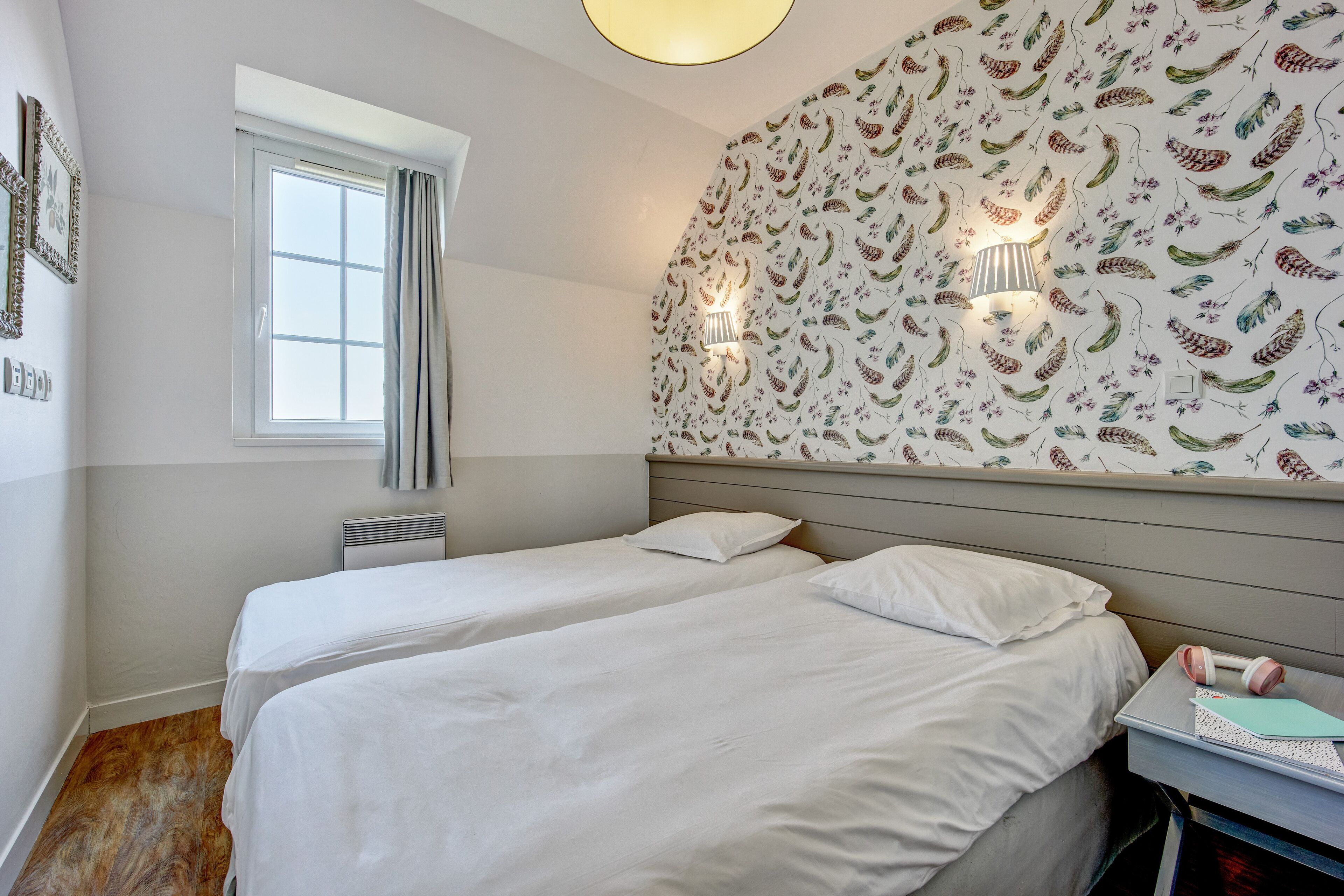 A bedroom with two beds, a window, a wall-mounted light fixture, and a wall with a decorative pattern.