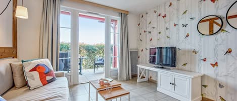 A modern living room with a sofa, a coffee table, a TV on a white cabinet, and a balcony with curtains.