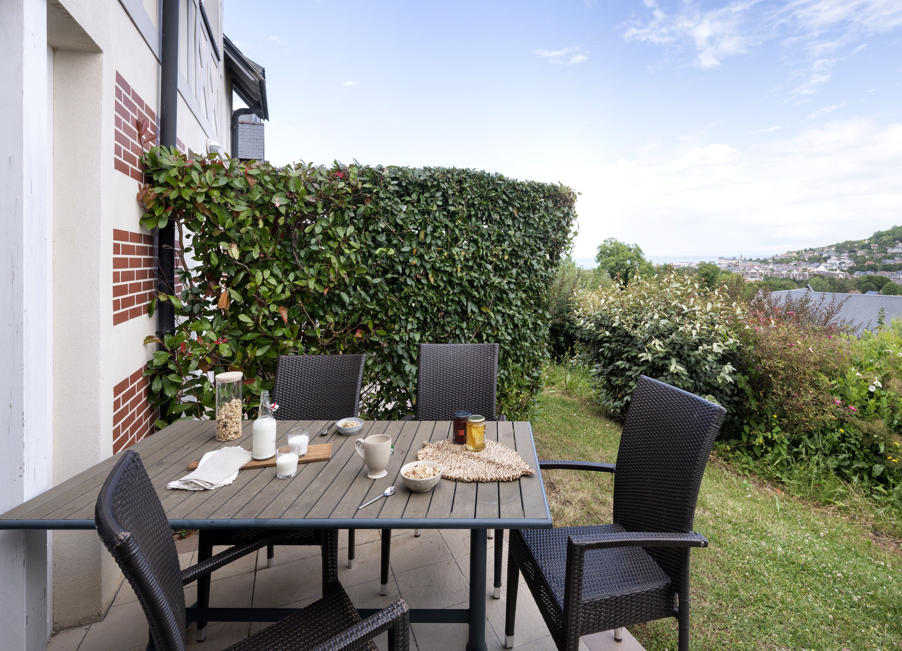 Un patio al aire libre con una mesa preparada para dos personas, rodeado de sillas de mimbre y una vista de la ciudad de fondo.