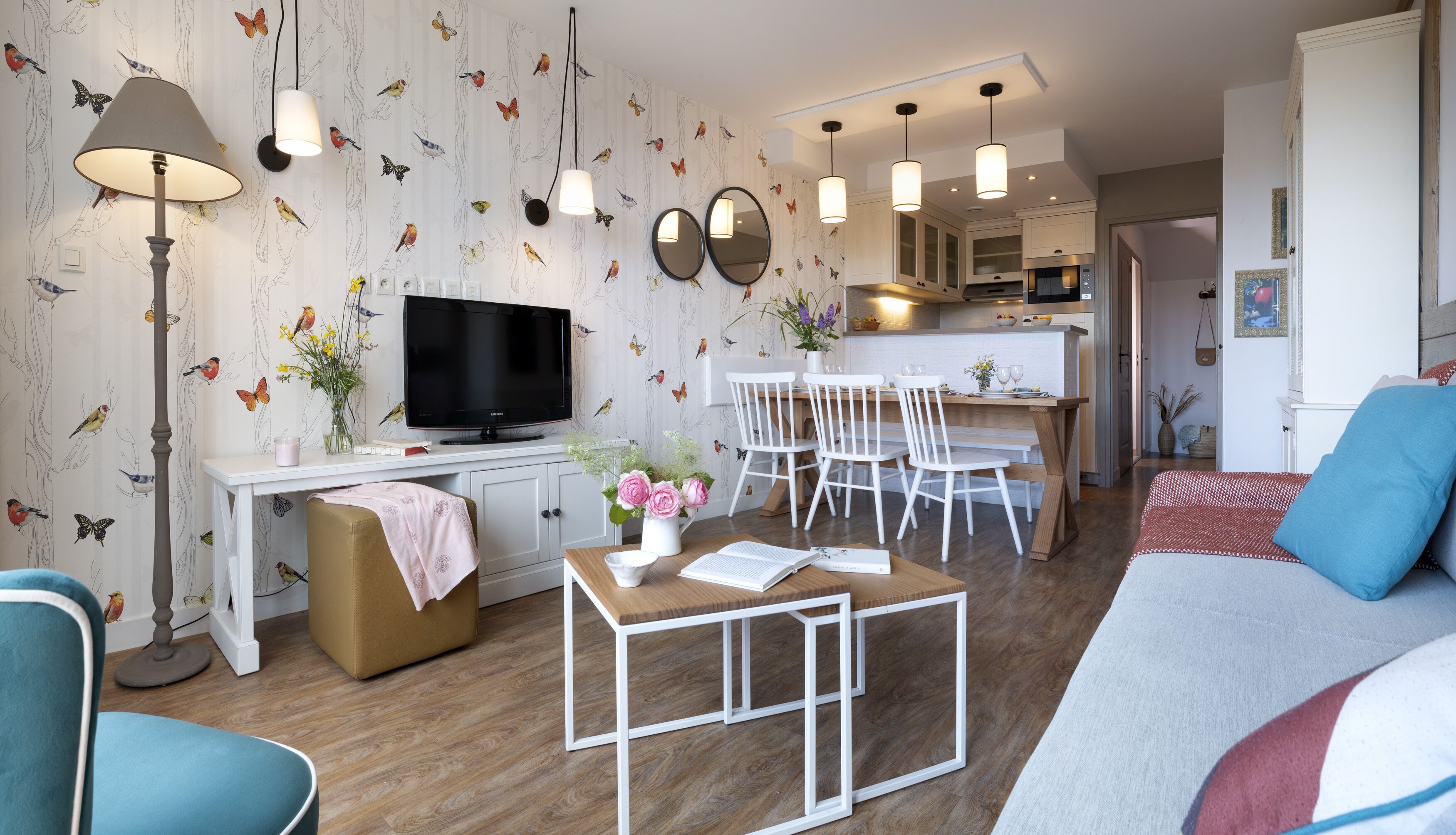 Una sala de estar moderna con un televisor de pantalla plana, un comedor con sillas blancas, una cocina al fondo y una zona de estar acogedora con un sofá y sillones.