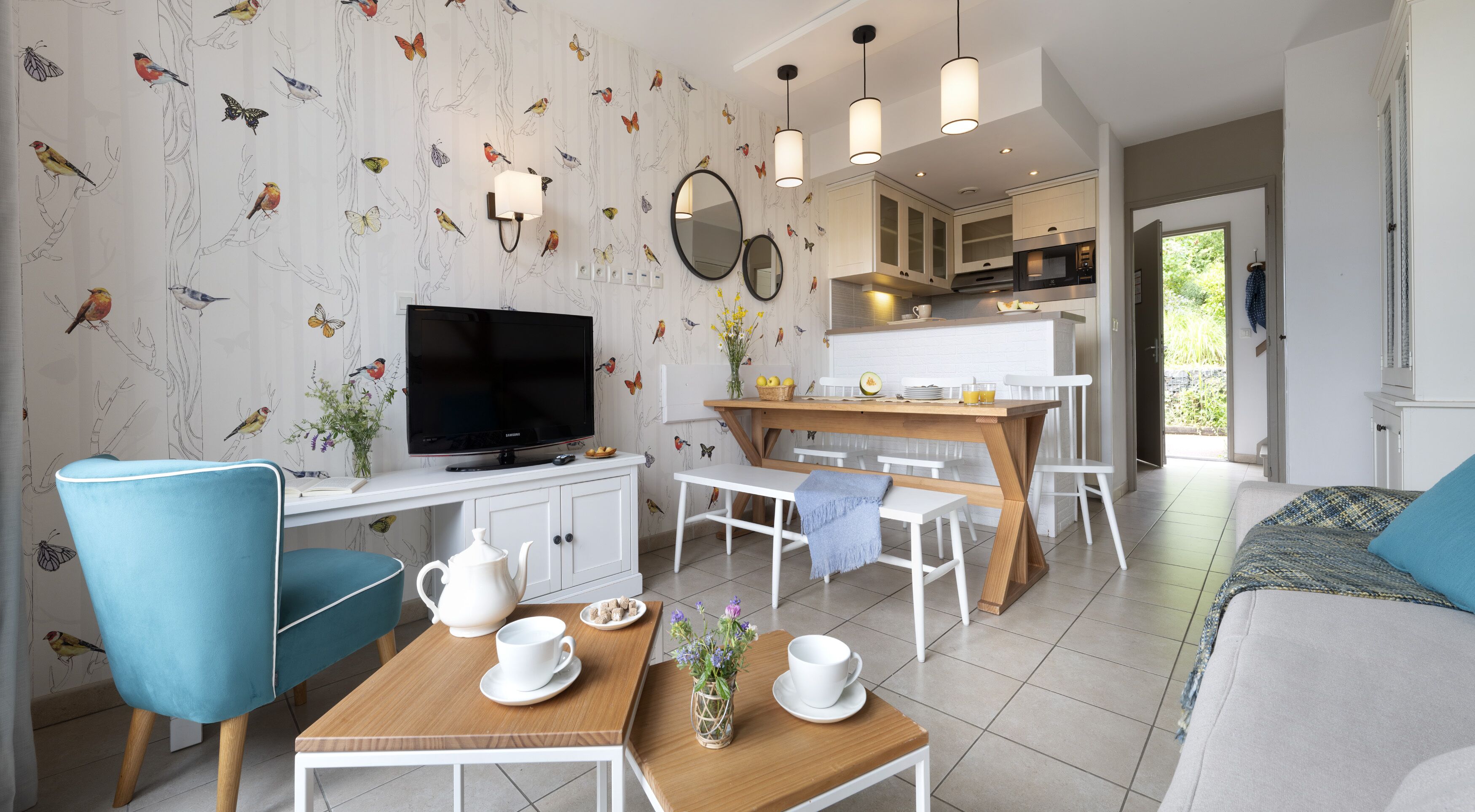 Een moderne keuken met witte kasten, een eethoek met een houten tafel en stoelen, een tv op een witte standaard, en een zithoek met een blauwe fauteuil en een bank.