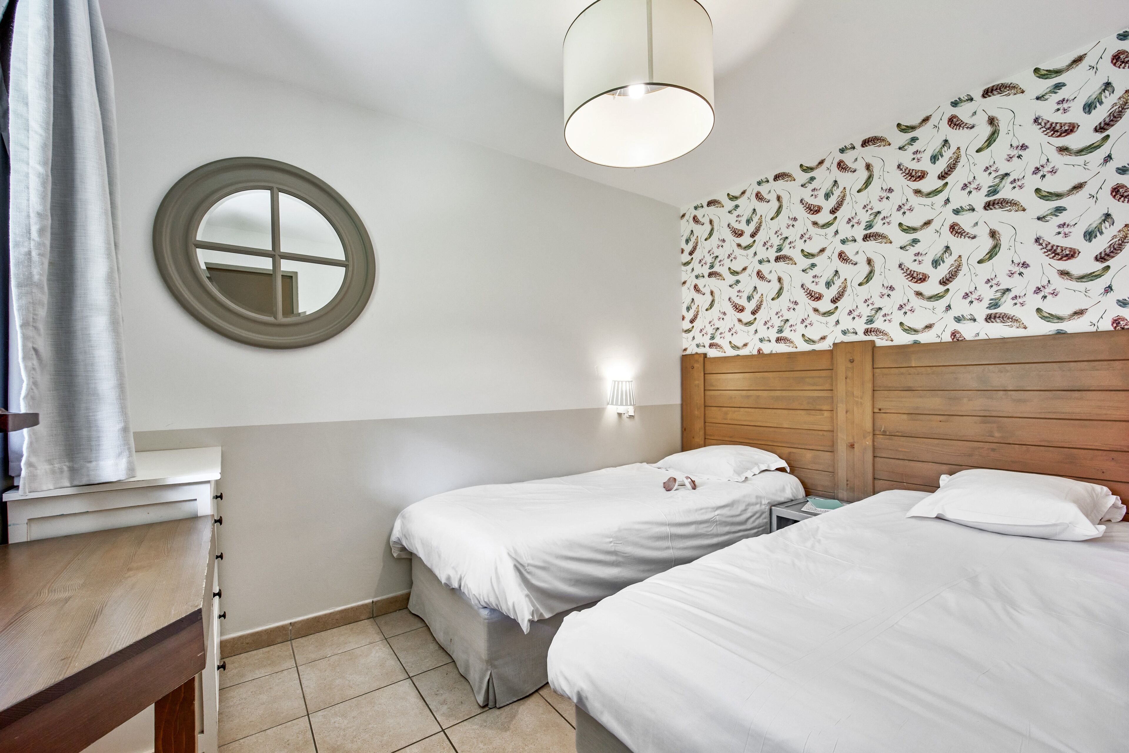 Une chambre avec deux lits simples, une tête de lit en bois, un miroir rond et un luminaire fixé au mur.
