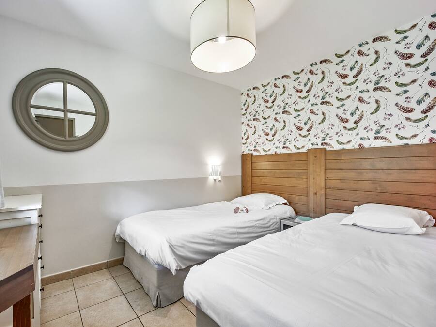 Une chambre avec deux lits simples, une tête de lit en bois, un miroir rond et un luminaire fixé au mur.
