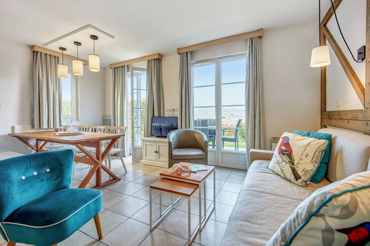 Ein modernes Wohnzimmer mit einer Couch, einem blauen Sessel, einem Holztisch, einem Esstisch mit Stühlen und einem Fernseher.