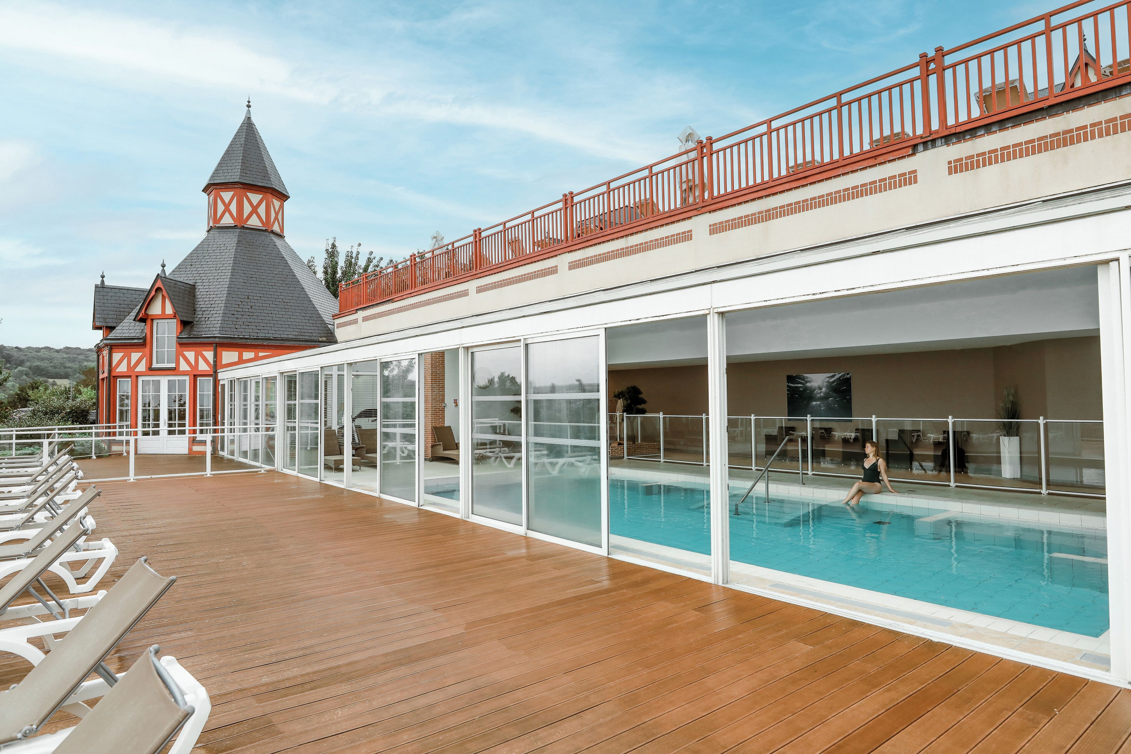 A poolside area with a wooden deck, glass-enclosed pool, and a building with a red and white facade.