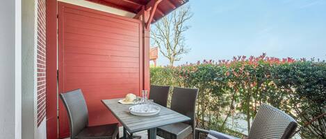 A balcony with a red door, two wicker chairs, a table with a tray, and a clear view of the sky and foliage.