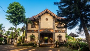 Un bâtiment traditionnel doté d’une entrée centrale, entouré d’une végétation luxuriante et bordé d’un chemin pavé.