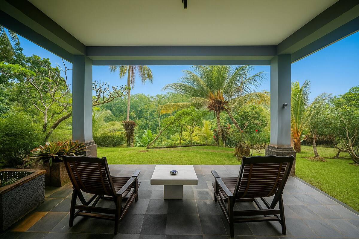 A covered patio with two wooden chairs and a table, overlooking a lush garden with palm trees and a clear blue sky.