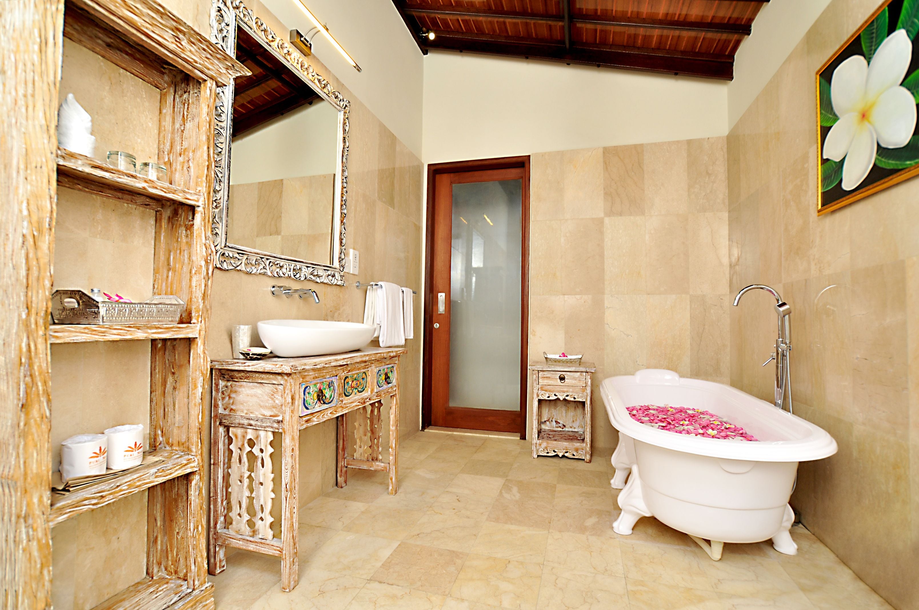 Ein Badezimmer mit einer freistehenden Badewanne, die mit Rosenblüten gefüllt ist, einem großen Spiegel und einem Holzwaschtisch mit dekorativer Schrankwand.