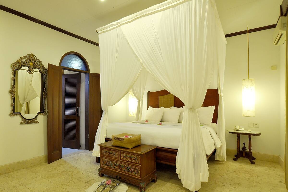 A bedroom with a four-poster bed, a wooden chest of drawers, a bedside table, a mirror, and a door leading to another room.
