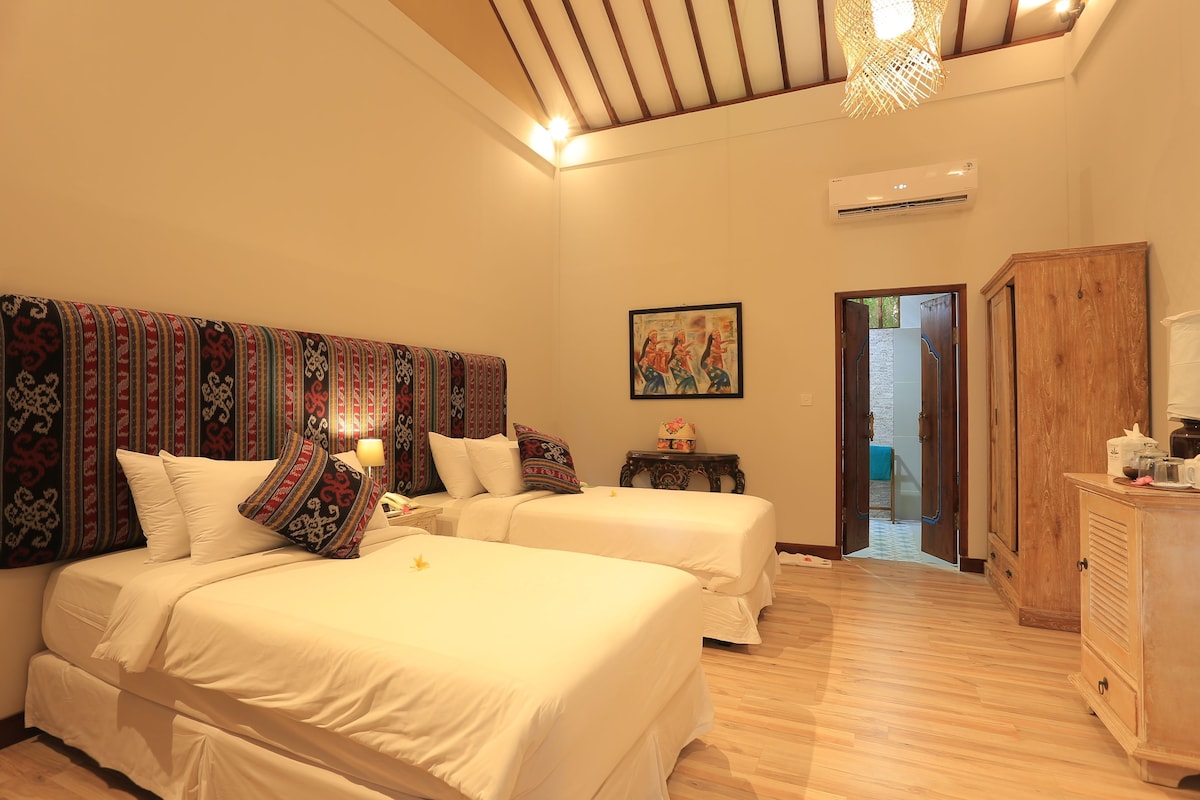 A hotel room with two beds, a wooden wardrobe, a small table, and a painting on the wall.