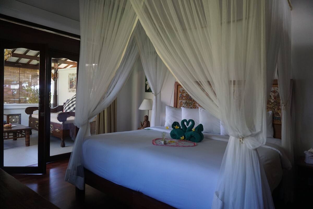 A bedroom with a canopy bed, a small table with a vase, and a view of an outdoor area.