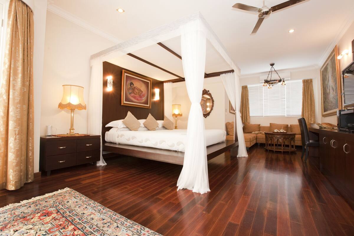 A bedroom with a four-poster bed, a nightstand, a lamp, a sofa, a dining table, and a television.