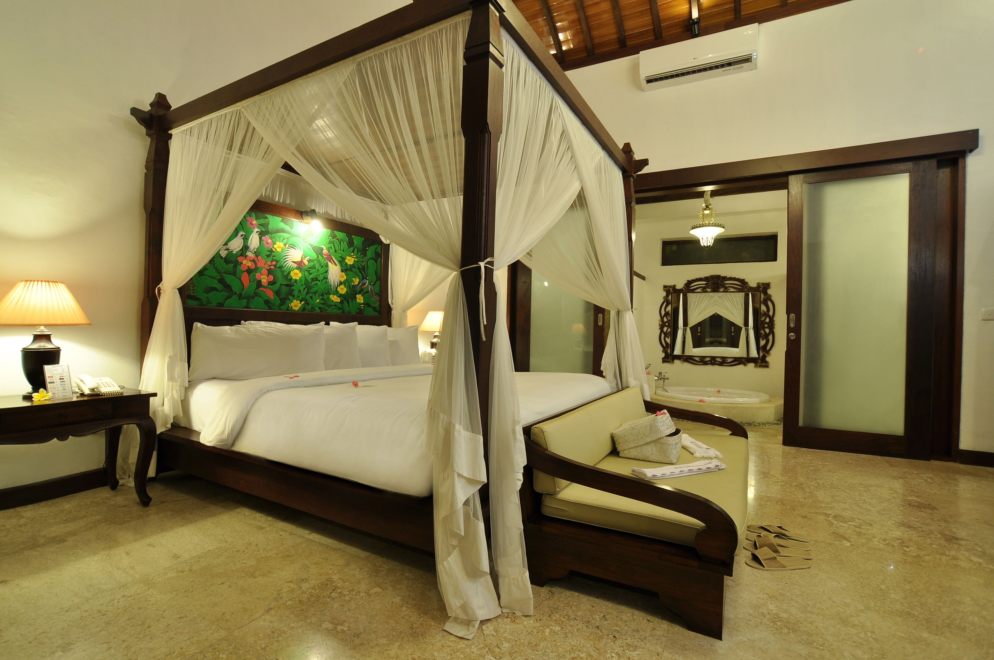 A four-poster bed with a canopy, a bedside table with a lamp, a nightstand, a sofa, a mirror, and a bathroom with a sink.