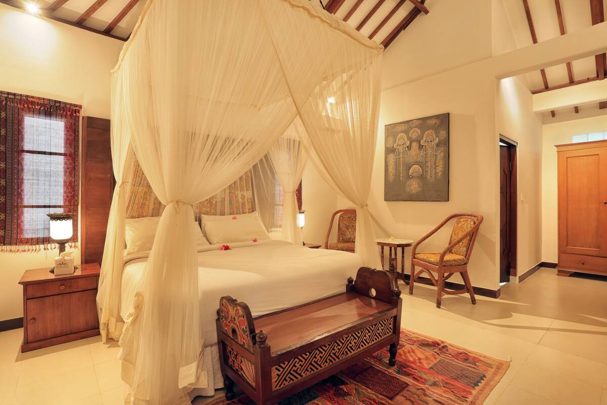 A bedroom with a four-poster bed, a nightstand, a chair, and a painting on the wall.