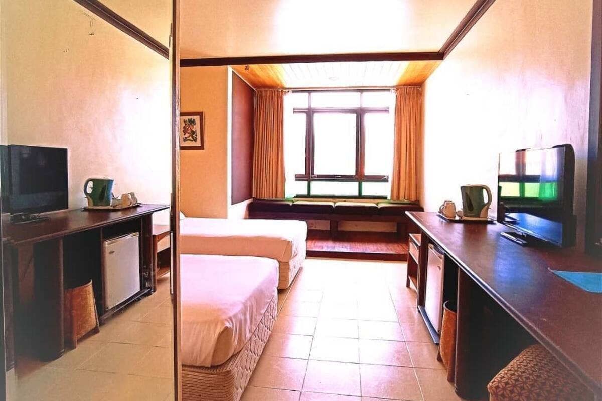 A hotel room with a bed, a desk with a television, a window with curtains, and a small kitchenette area.