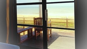 A view from a room with a large window overlooking a wooden deck and a beach.