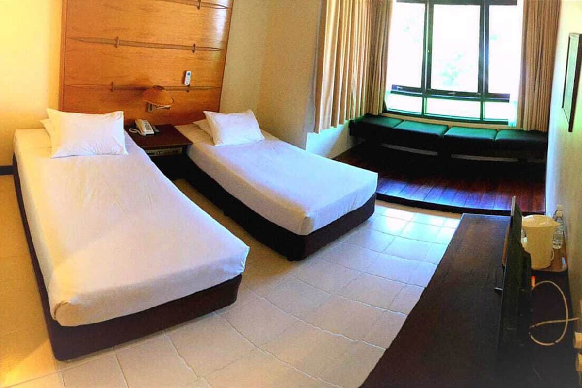 A hotel room with two single beds, a wooden headboard, a small desk with a telephone, and a window with curtains.