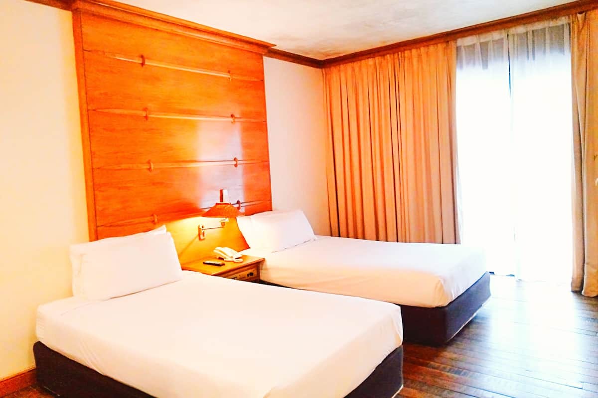 A hotel room with two beds, a wooden headboard, a bedside table with a lamp, and a window with curtains.