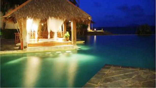 A thatched-roof structure with a pool, likely a resort or hotel, with a view of the ocean at dusk.