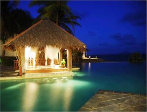 A thatched-roof structure with a pool, likely a resort or hotel, with a view of the ocean at dusk.