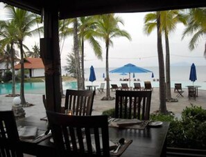 An outdoor seating area with tables and chairs, overlooking a pool and palm trees.