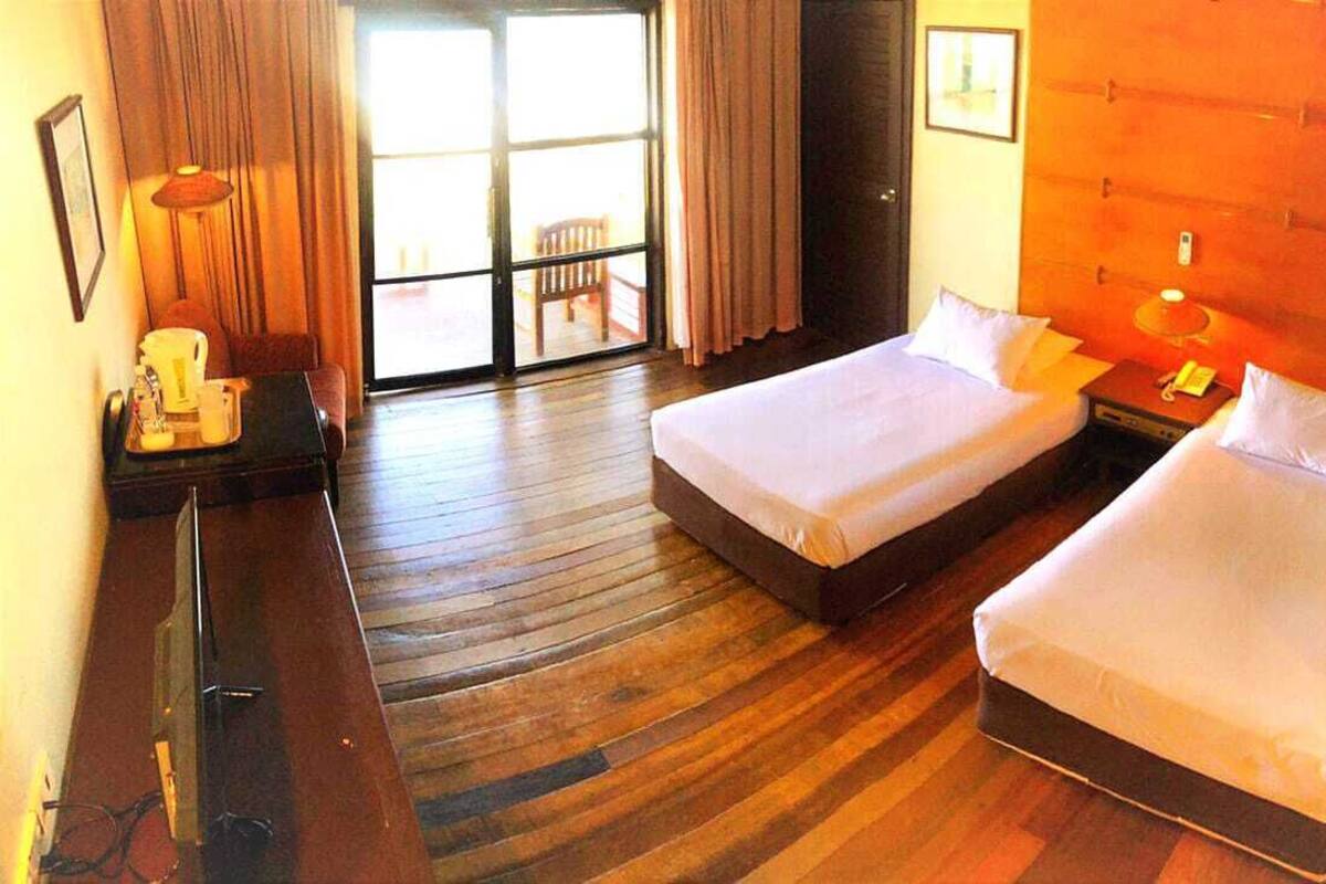 A hotel room with two single beds, a wooden floor, a desk with a lamp, and a view of the outdoors through a sliding door.