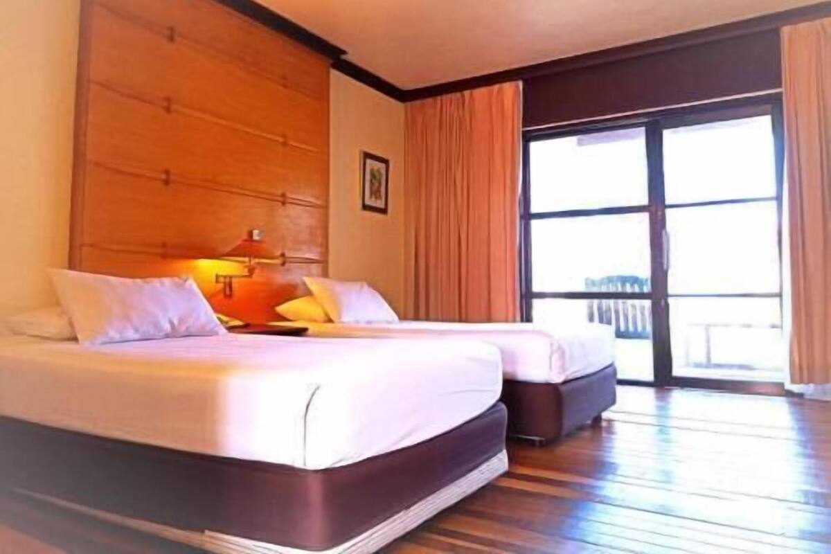 A hotel room with a large bed, a bedside table, a window with curtains, and a wooden floor.