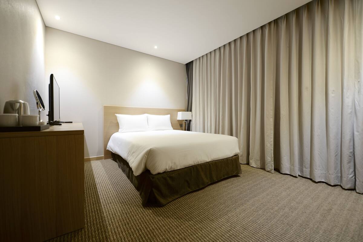 Een hotelkamer met een bed, een bureau, een lamp en een groot raam met gordijnen.