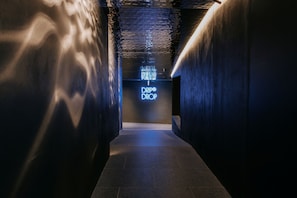 Ein Korridor mit einer spiegelnden Decke und einem leuchtenden Neon-Schild mit der Aufschrift 'DRIP DROP' in der Ferne.